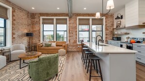 Une cuisine moderne avec un mur en briques, un grand îlot central doté de tabourets et un coin salon comprenant un canapé et des fauteuils.
