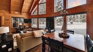 A cozy cabin interior with a wooden ceiling, large windows, a fireplace, and a dining area. - McCall A-Frame on Golf Course, 5 Minutes from Town + Huge Yard, Hot Tub & 7 Beds (McCall)