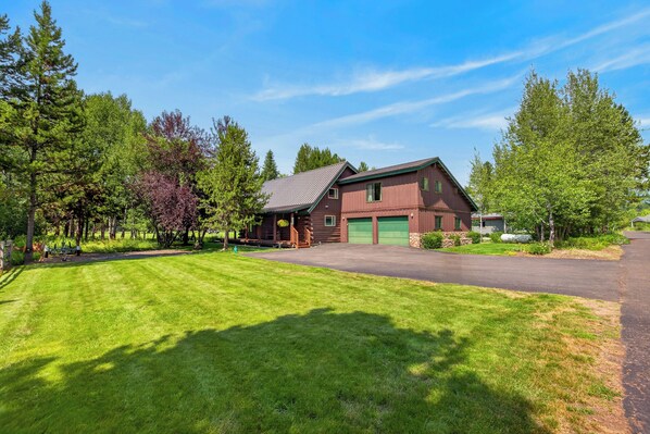 A two-story log cabin with a green garage, surrounded by a well-maintained lawn and trees.