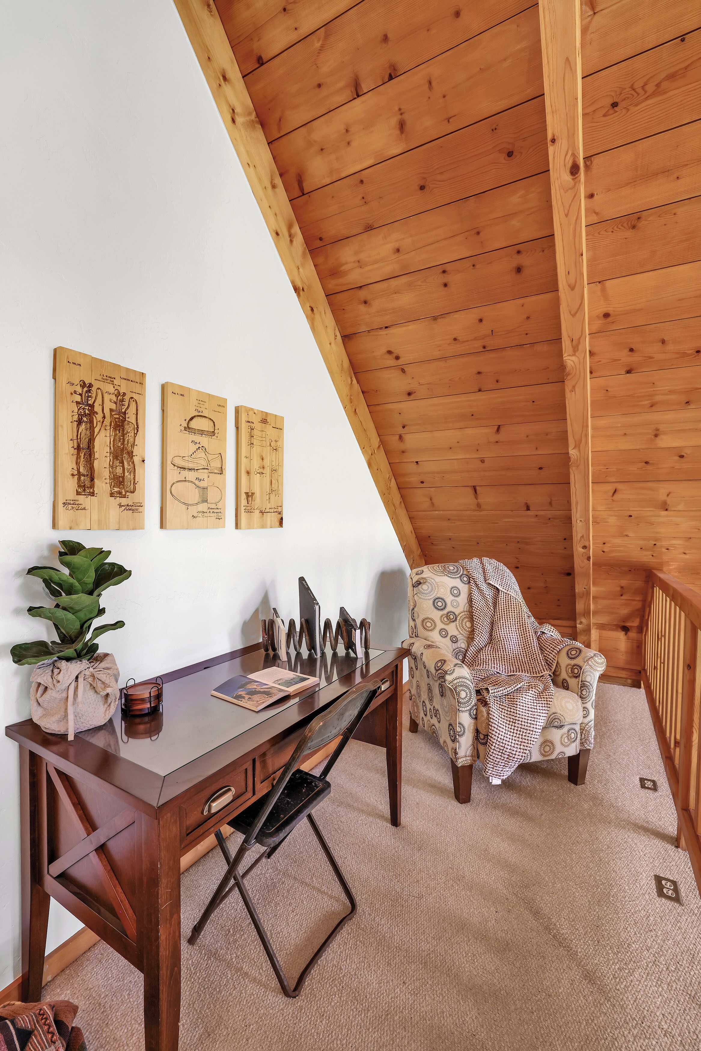 A cozy room with a wooden ceiling, a desk with a chair, and three framed drawings on the wall.
