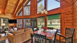 A wooden cabin interior with a dining area, sofa, and large windows offering a view of the outdoors. - McCall A-Frame on Golf Course, 5 Minutes from Town + Huge Yard, Hot Tub & 7 Beds (McCall)