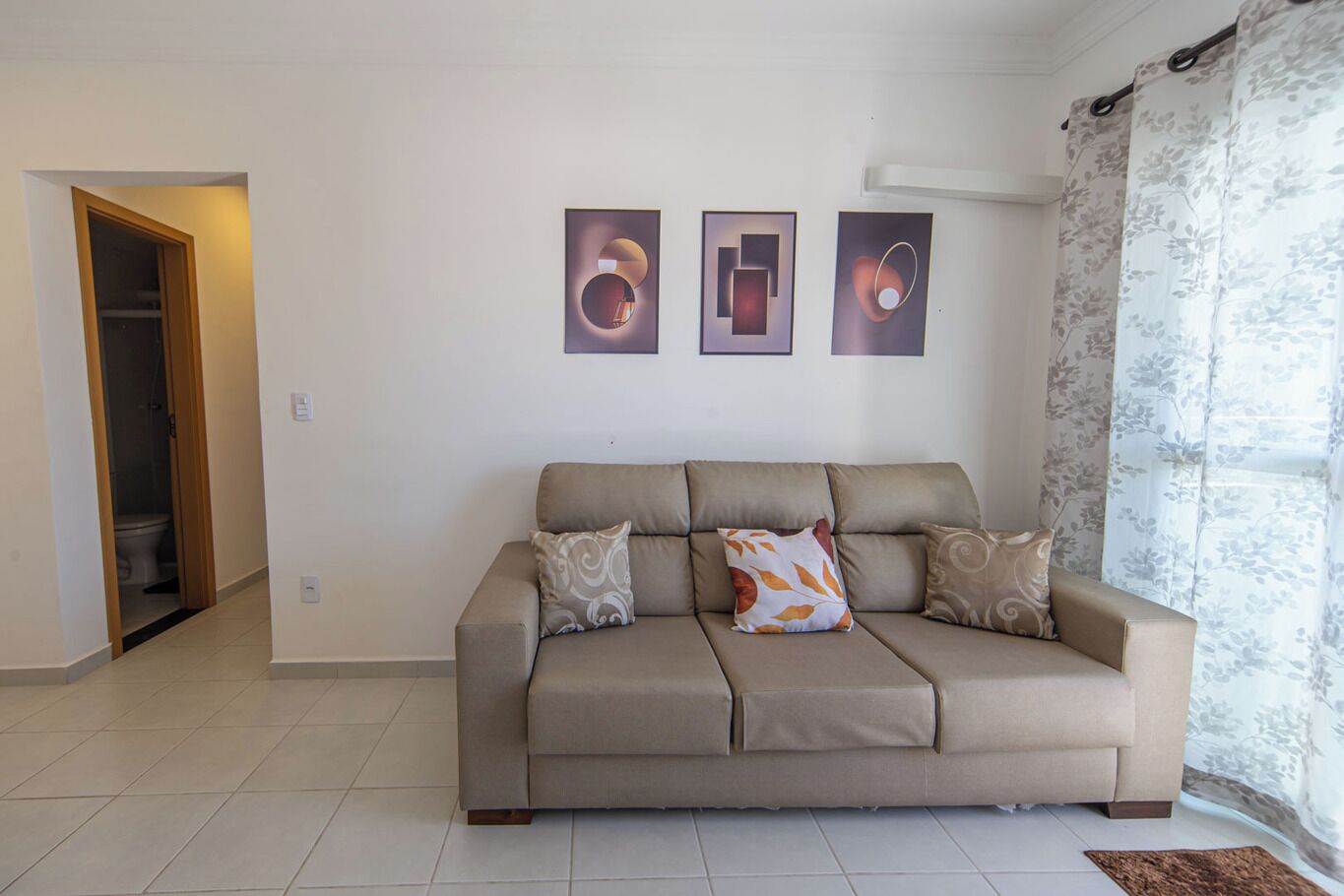 Ein Wohnzimmer mit einem beigen Sofa, drei gerahmten Kunstwerken und einem gemusterten Vorhang.