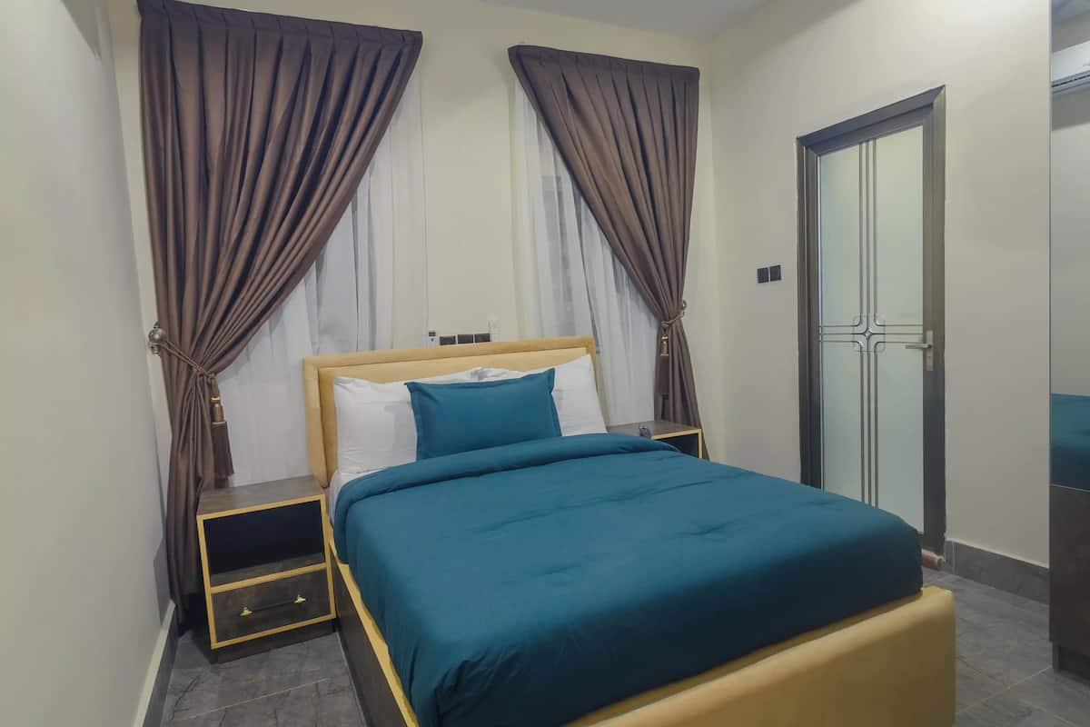 A neatly made bed with a blue bedspread and white pillows, flanked by two nightstands with drawers, in a room with a door and curtains.