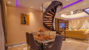 A modern interior with a spiral staircase, a dining area with a wooden table and chairs, a sofa, and a TV in the background. - Quads Villa (Accra)