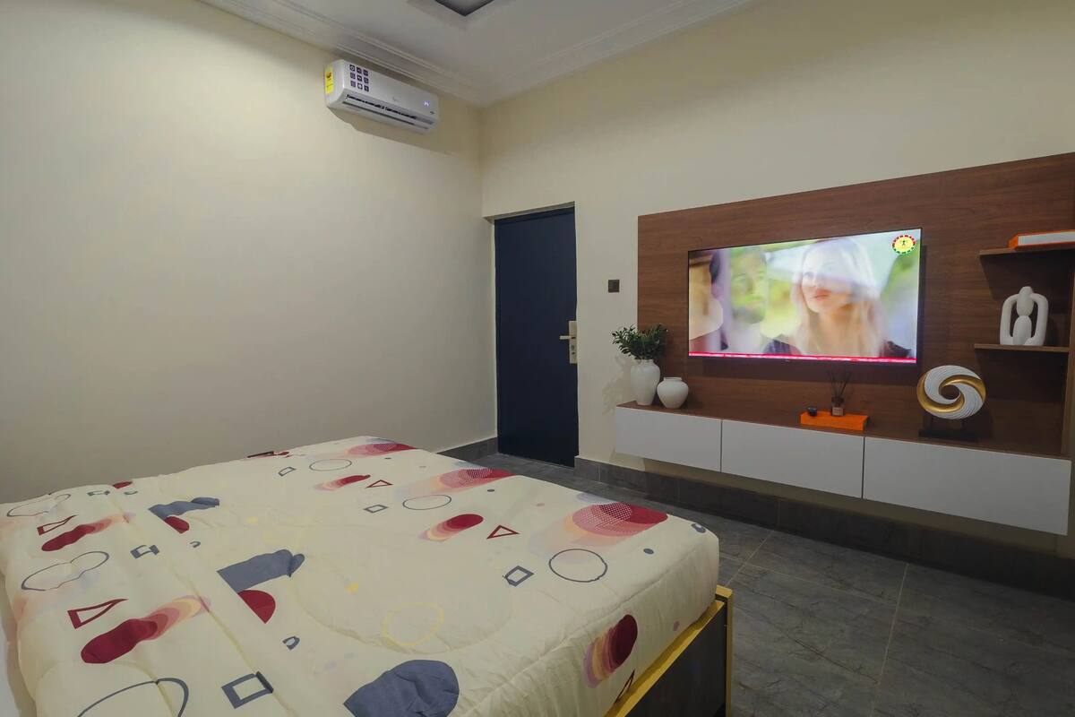 A hotel room with a bed, a TV mounted on the wall, a door, and a shelf with decorative items.