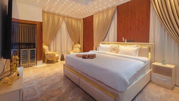 A spacious bedroom with a large bed, a gold-framed armchair, a television, and a nightstand. - Quads Villa (Accra)