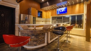 A modern bar area with a stone counter, two red bar stools, and a television screen displaying a news channel. - Quads Villa (Accra)