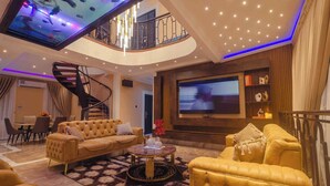 A spacious living area with a large flat-screen TV, a comfortable sofa set, a round coffee table, and a spiral staircase leading to an upper level. - Quads Villa (Accra)