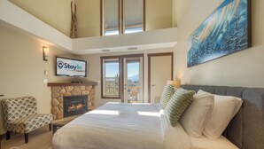 Un'ampia camera da letto con un letto grande, un camino in pietra, una televisione e un balcone con vista sulle montagne.