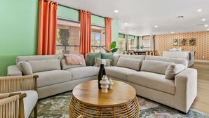 A living room with a sectional sofa, a round wooden coffee table, and a dining area with a white cabinet and patterned wallpaper. - Family Fun Oasis: Heated Pool, Pickleball, Basketball, Mini Golf & Game Room (Clearwater)