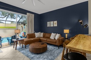A modern living room with a sectional sofa, a round coffee table, a dining table with chairs, and a poolside area with a table and chairs. - 3BR Pool Villa w Resort Access, Disney Area (Kissimmee)