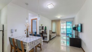 A modern living room with a dining area, a sofa, a TV mounted on the wall, and a large window with curtains. - Your Exclusive Stay in a Building With a Pool Just Steps From the sea - WJA (Salvador)