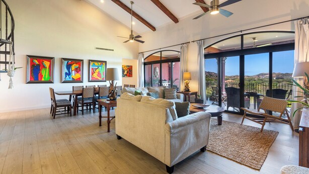 A spacious living room with a high ceiling, wooden beams, a dining area, and a balcony with a view.