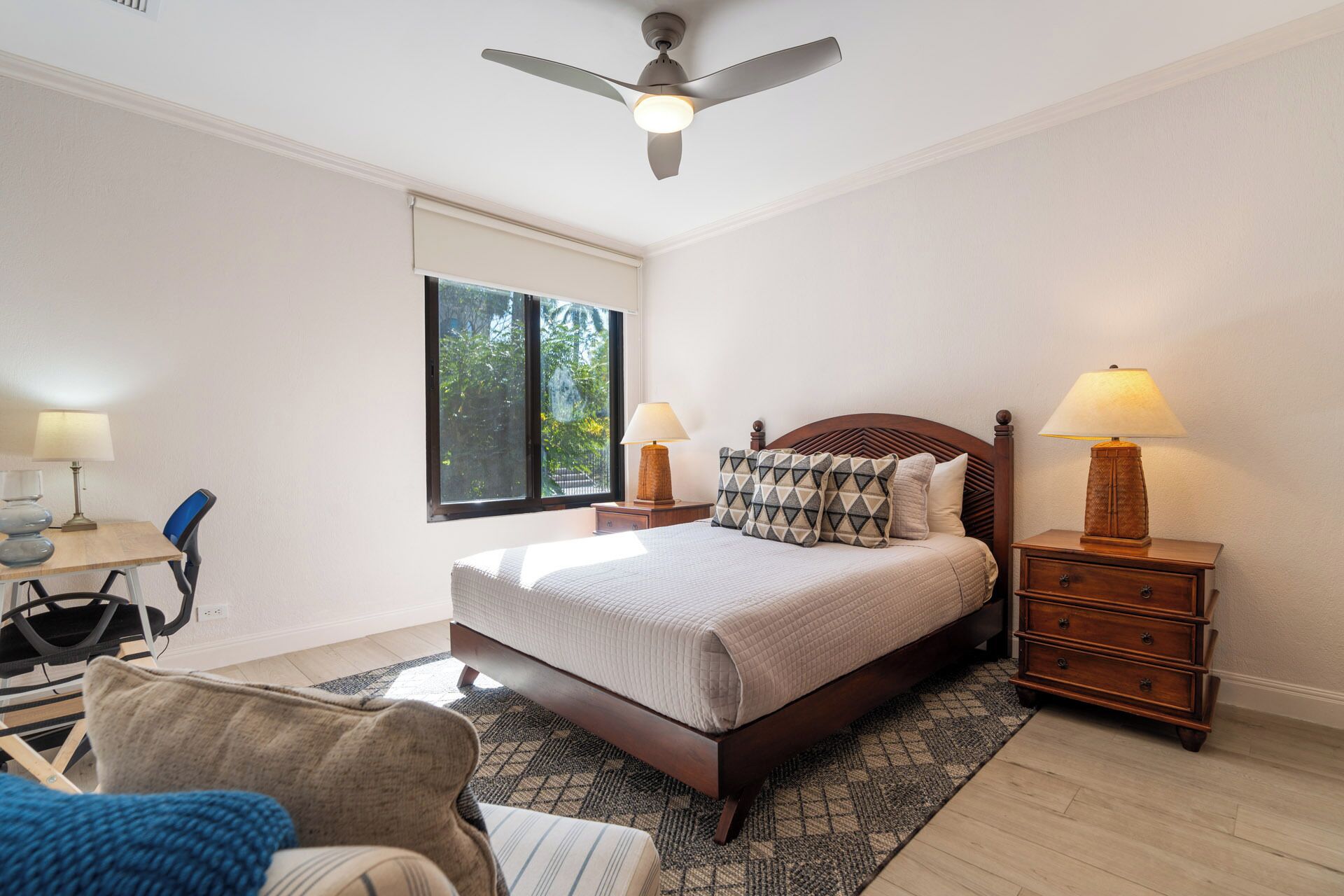 A bedroom with a bed, two bedside tables, a chair, a desk, a window, and a ceiling fan.