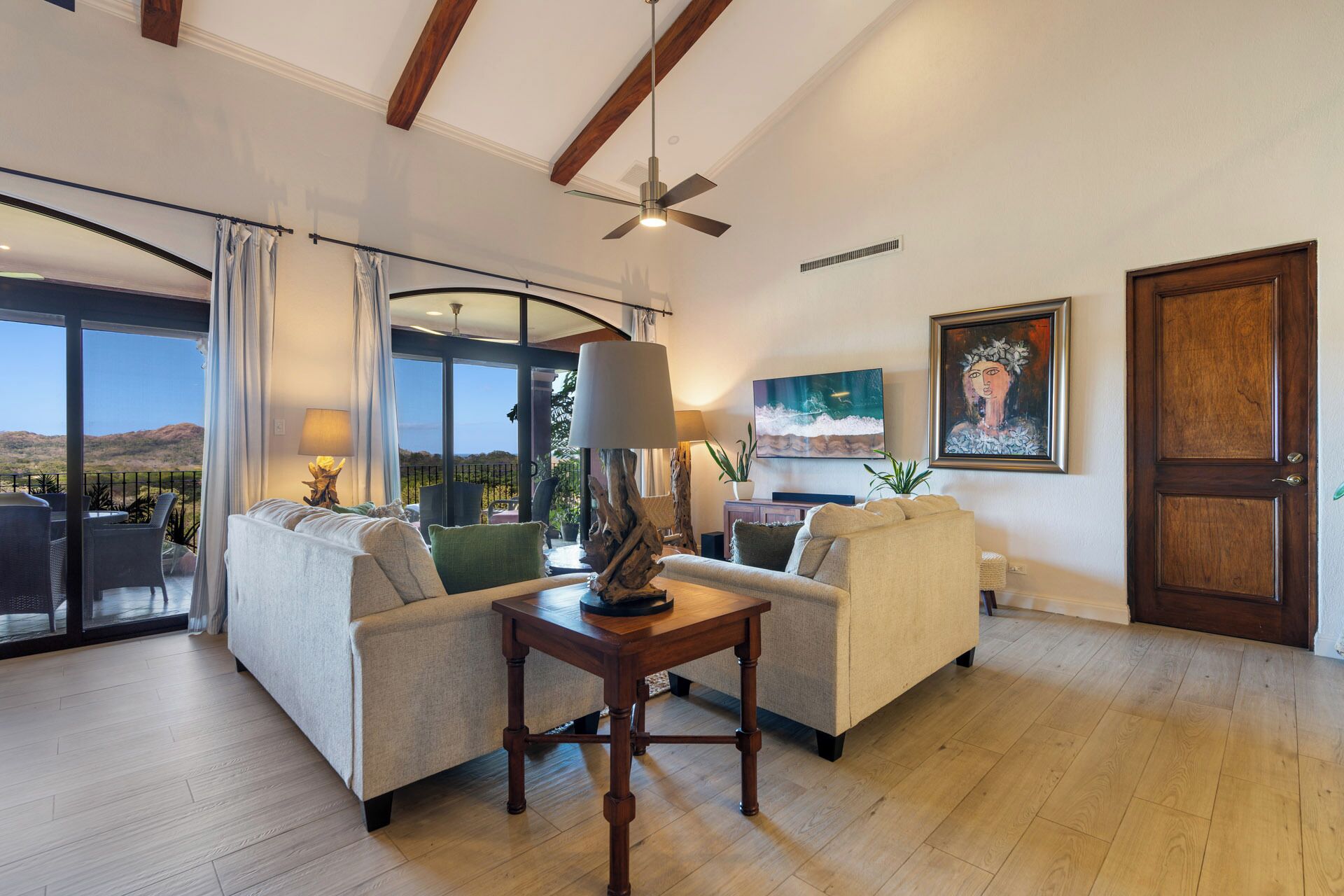 A spacious living room with a large sliding glass door leading to a balcony, a ceiling fan, a flat-screen TV, and a painting on the wall.