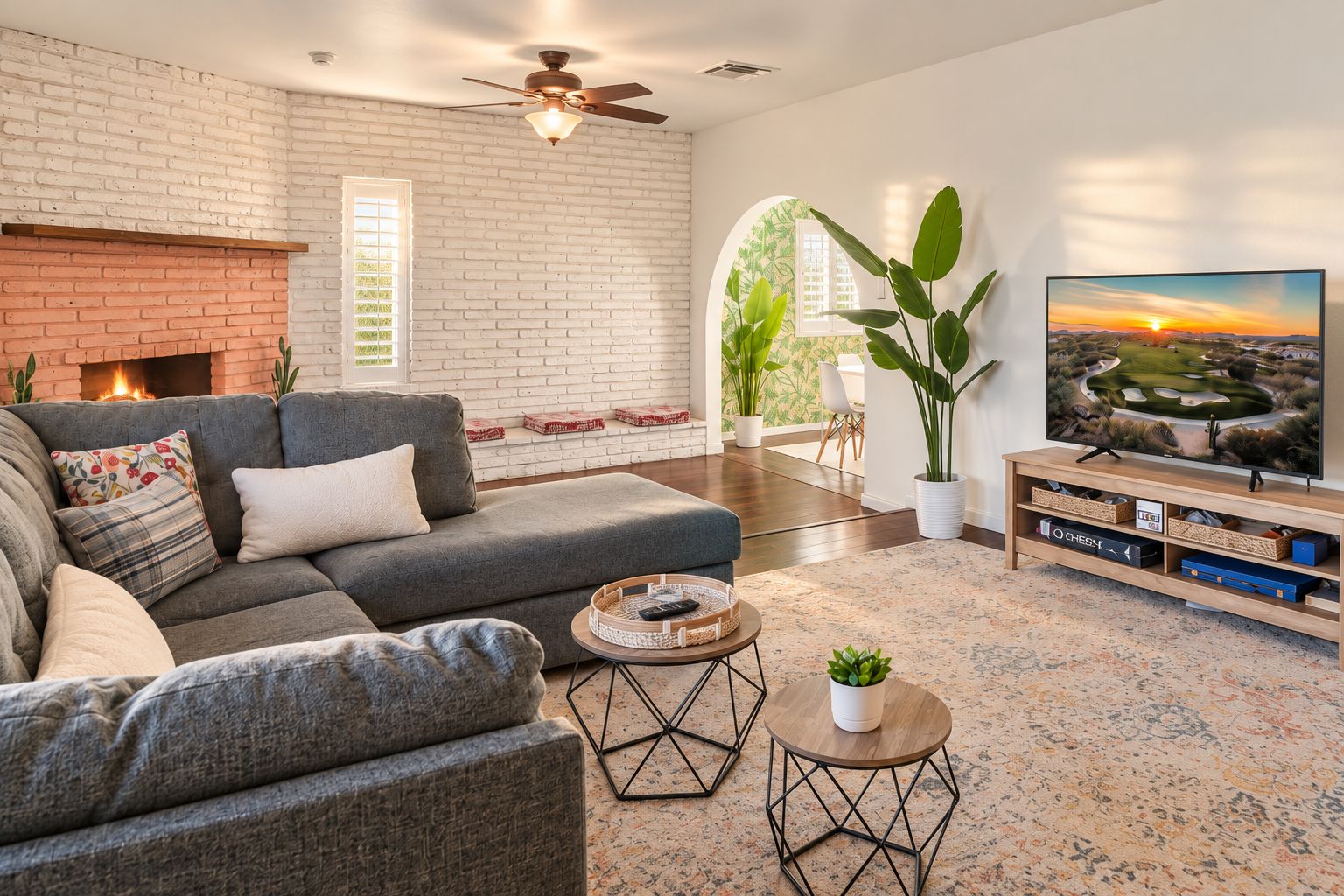 A living room with a brick fireplace, a grey sofa, a television, and a large window with a view of the sunset.
