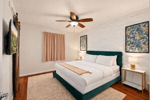 Une chambre à coucher avec un lit, un ventilateur de plafond, une table de chevet avec une lampe, une fenêtre avec des rideaux et des œuvres d’art accrochées aux murs.