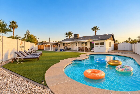 A backyard with a swimming pool, lounge chairs, and a covered patio area.