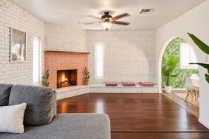 A spacious living room with a brick fireplace, a ceiling fan, and a large window. - The Pine Desert Retreat - Scottsdale's Ultimate Getaway (Scottsdale)