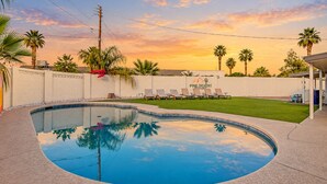 A swimming pool with a curved edge, surrounded by a white wall and palm trees. There are lounge chairs and a covered area with a sign that reads 'Pine, Desert Resort.'