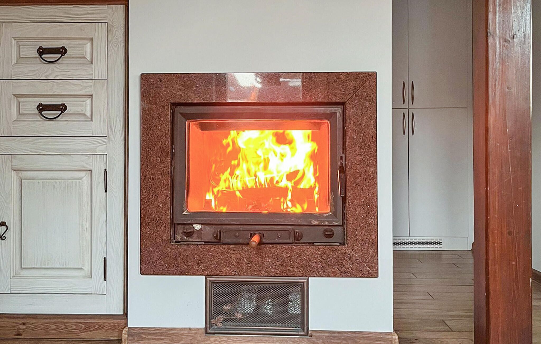 A wood-burning stove with a fire burning inside, mounted on a wall with a white cabinet to the left and a wardrobe to the right.