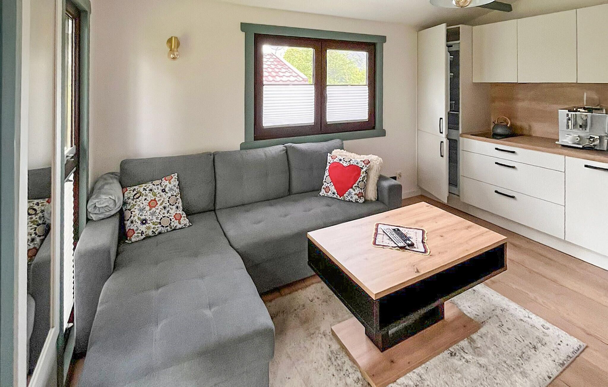 Un espace de vie compact comprenant un canapé gris, une table basse en bois, une fenêtre avec des stores et une kitchenette dotée de meubles blancs.