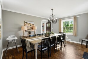 A dining room with a wooden table, black chairs, a chandelier, a window with curtains, and a white chair. - Hot Tub • Fenced Yard • Perfect Family Retreat Near Chagrin Falls (Geauga County)