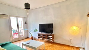 Een woonkamer met een groene bank, een witte salontafel en een televisie op een houten dressoir. Op de vloer ligt een tapijt met een patroon en er staat een potplant bij het raam.