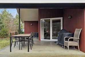 An outdoor deck with a table and chairs, a covered grill, and a sliding glass door leading to a covered patio area. - Peaceful cottage near beach and nature (Hadsund)