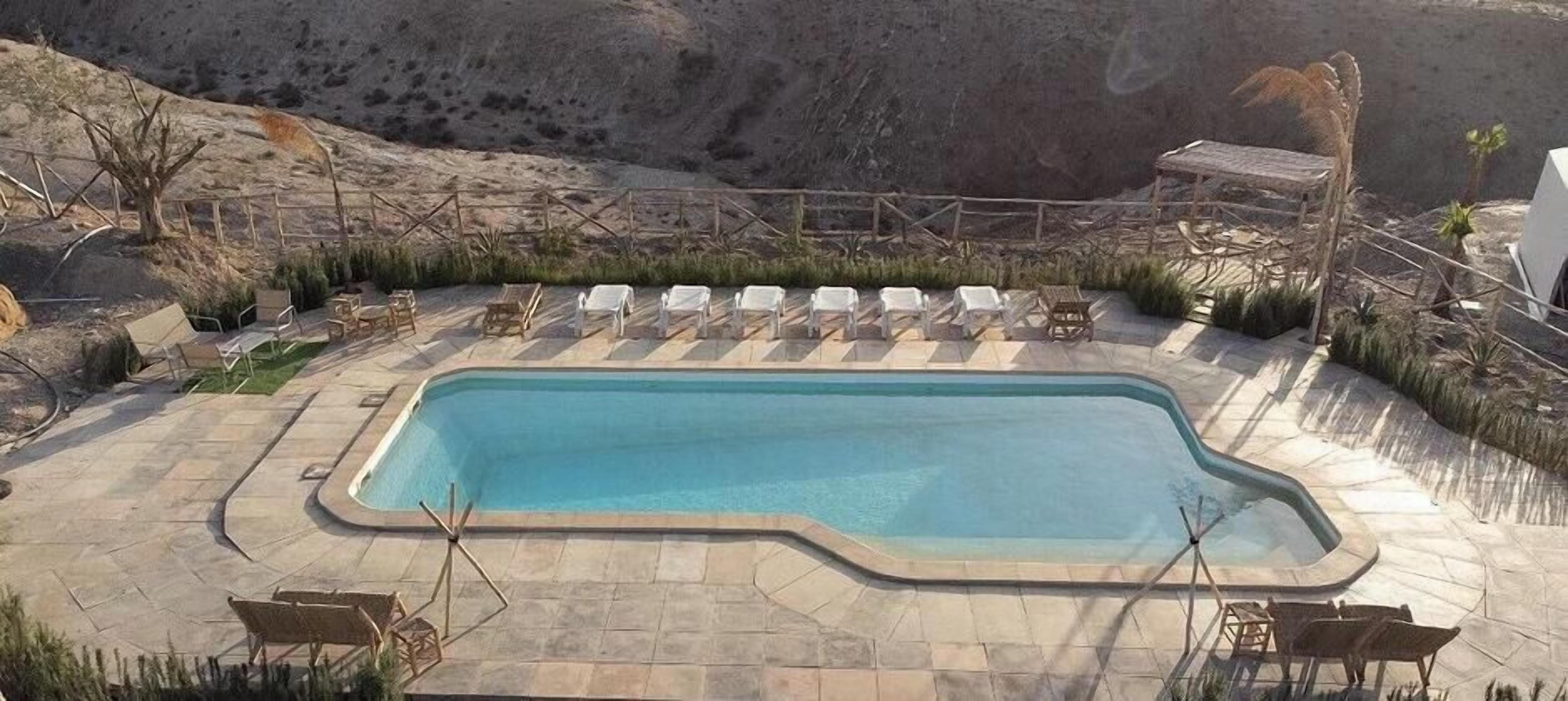 Piscina rectangular con solárium alicatado, rodeada de sillones blancos y una valla de madera.