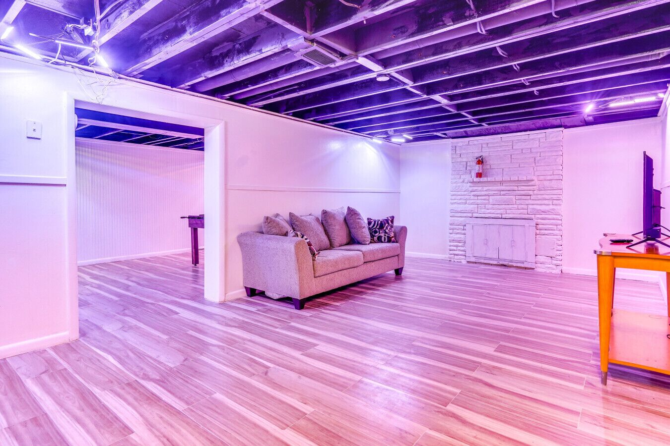 A spacious room with a wooden floor, a grey sofa with patterned cushions, and a white brick wall.