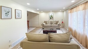 A living room with a beige sofa, a coffee table, and a couch. There are two framed botanical prints on the wall, a potted plant on a side table, and a window with sheer curtains.