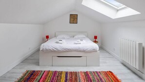 Een slaapkamer met dakraam, witte muren, een bed met witte beddengoed, een kleurrijk gestreept tapijt en een rode lamp aan elke kant van het bed.