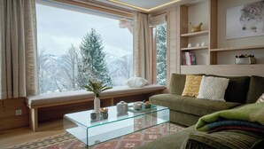 Un salon chaleureux avec une grande fenêtre, une table basse en verre, un canapé et une bibliothèque.