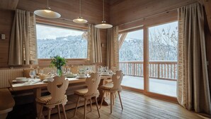 Une salle à manger en bois avec une table dressée pour quatre personnes, de grandes fenêtres avec des rideaux et une vue sur des arbres couverts de neige.