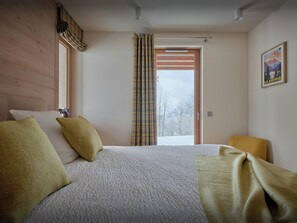 A bedroom with a bed, a chair, a window with curtains, a door, and a wall with a framed picture.