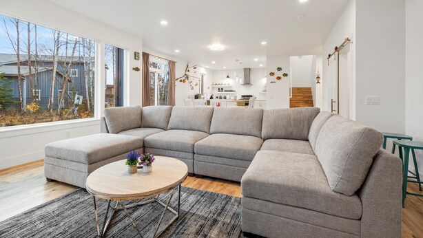 A modern living room with a grey sectional sofa, a round wooden coffee table, and a view of the kitchen and backyard through large windows. - ANCHORAGE HOUSE WITH PEACEFUL PLACE (Anchorage)
