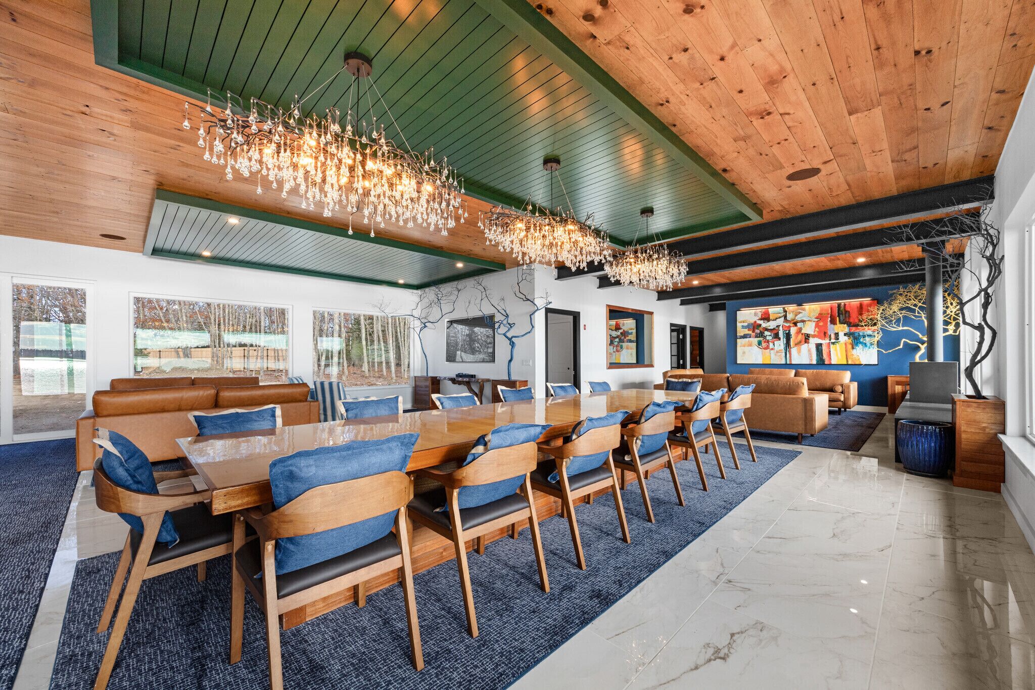 A spacious room with a long wooden table and blue upholstered chairs, a chandelier, and a large window with a view of trees.