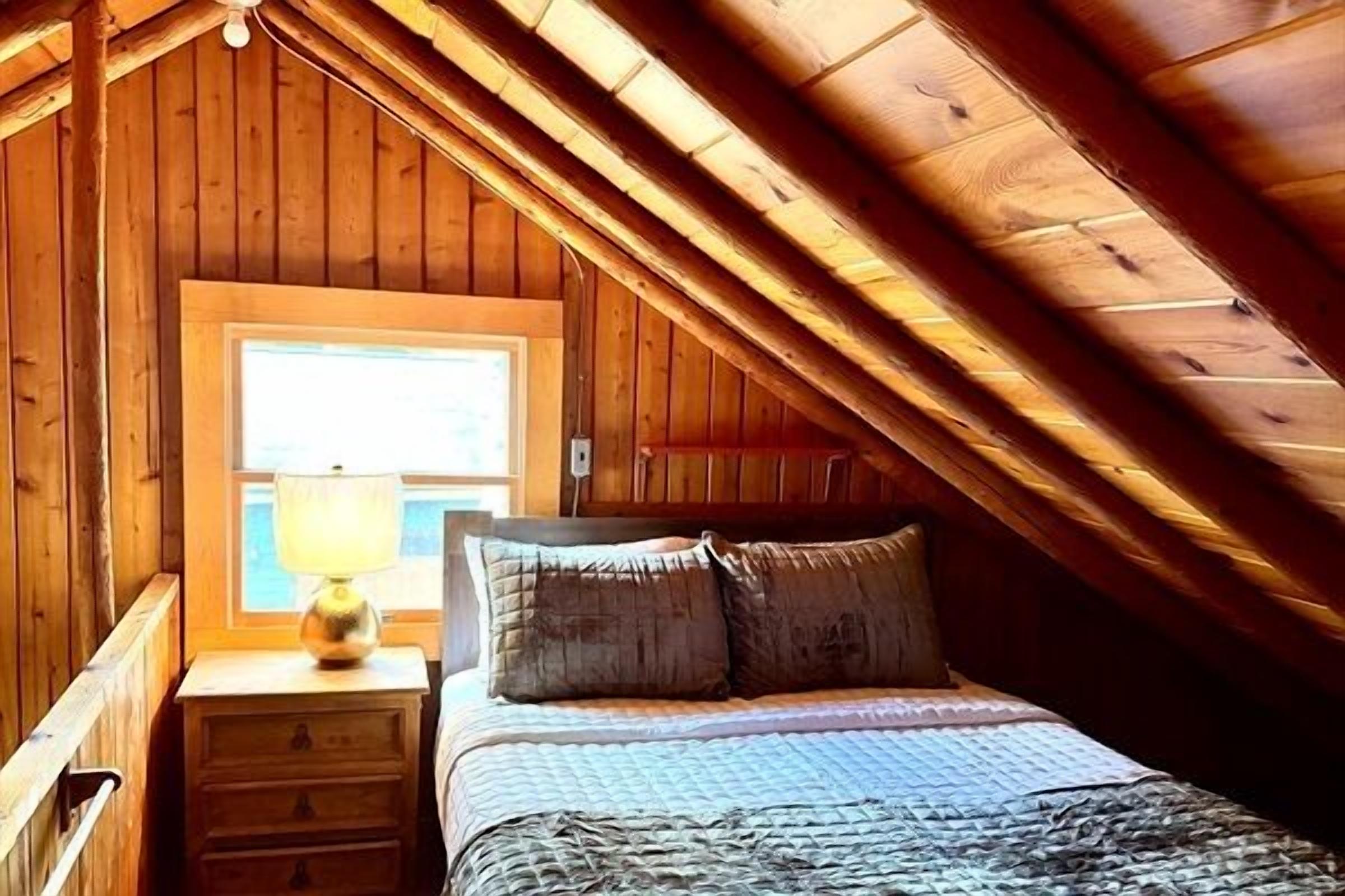 A bedroom with a wooden ceiling, a window, a bedside table with a lamp, and a bed with pillows.