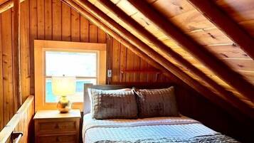 A bedroom with a wooden ceiling, a window, a bedside table with a lamp, and a bed with pillows.