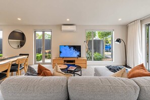 Ein modernes Wohnzimmer mit einem großen Flachbildfernseher, einem Essbereich mit einem Holztisch und Stühlen sowie großen Fenstern mit Blick ins Freie.