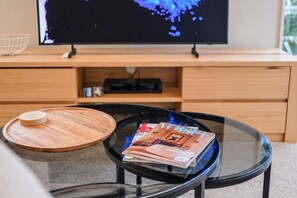 Ein Wohnzimmer mit Fernseher auf einem Holzboden, einem Glastisch mit Zeitschriften und einem Holztisch.