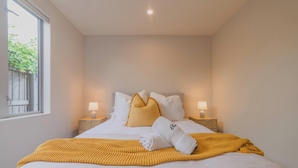 Ein ordentlich gemachtes Bett mit einer gelben Decke, weißen Kissen und einem zusammengerollten Handtuch. Auf den hölzernen Nachttischen stehen jeweils zwei Nachttischlampen.