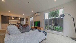 Ein modernes Wohnzimmer mit grauem Sofa, einem Flachbildfernseher und einem großen Fenster mit Blick auf einen Garten.