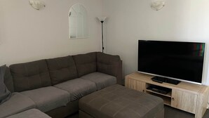 En stue med en hjørnesofa, et fladskærms-tv, en lampe og et træskab.