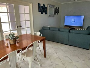 Una sala de estar con una mesa de comedor de madera, sillas blancas, un sofá gris, un televisor de pantalla plana y una pared con obras de arte enmarcadas.