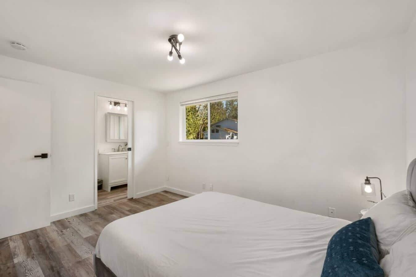 Quarto com uma cama grande, janela com vista, banheiro com pia e espelho, e luminária no teto.