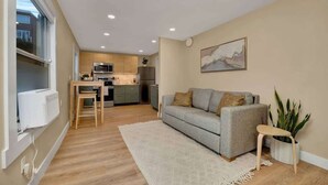 A modern living room with a grey sofa, a small table, and a plant. A kitchen with a microwave and refrigerator is visible in the background. - Westlake Nest | Minutes to DT | Quiet & Renovated! (Seattle)
