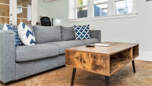 Un salon avec un canapé gris, deux coussins à motifs, une table basse en bois et une fenêtre avec des stores.