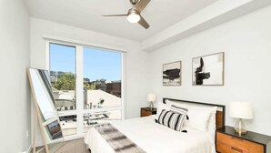 Ein modernes Schlafzimmer mit einem großen Bett, zwei Nachttischen, einem Deckenventilator und einem Blick auf die Stadt durch große Fenster.
